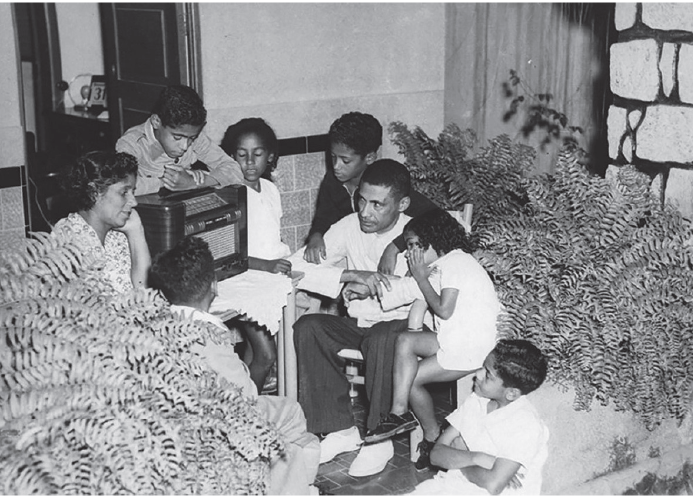 Imagem: Fotografia em preto e branco. Crianças sentadas ao redor de uma televisão pequena retangular. Há um homem e uma mulher sentados junto com as crianças. Fim da imagem.