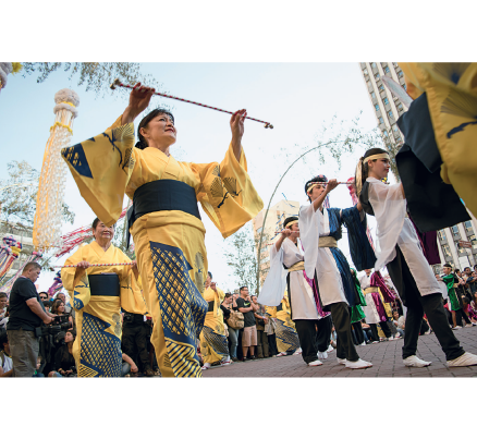 Imagem: Fotografia. Mulheres de quimono amarelo, segurando varinhas. Ao lado, fileira de meninos de quimono branco segurando varinhas. Ao redor, na rua, há pessoas observando. Fim da imagem.