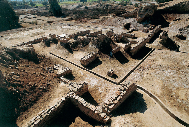 Imagem: Fotografia. Vista superior de campo de terra com demarcações no chão separando ruínas de sítios arqueológicos formados por muros de pedras incompletos.   Fim da imagem.