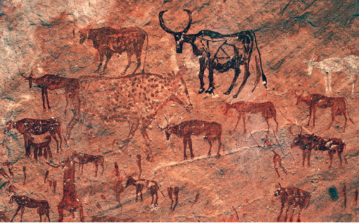 Imagem: Fotografia. Rocha com ilustração de gado em pinturas rupestres em branco, vermelho e preto. Há pessoas entre os animais.   Fim da imagem.