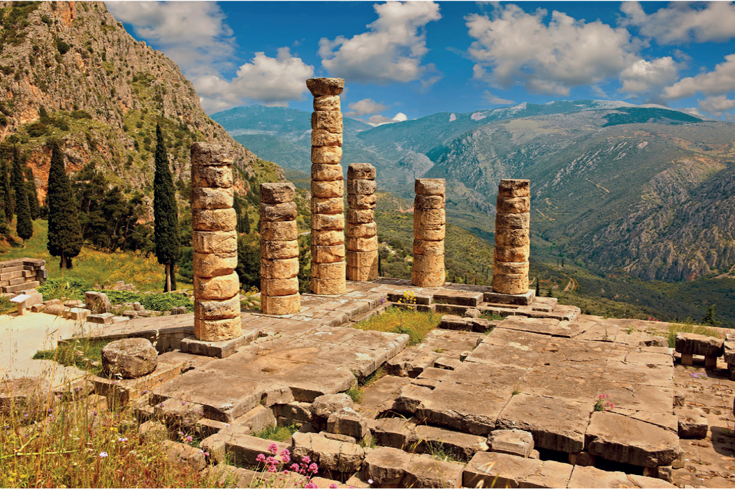 Imagem: Fotografia. Sitio arqueológico de um templo apenas com pedras no chão e colunas cilíndricas. Está sobre um monte com árvores e campo e ao fundo há montanhas.   Fim da imagem.