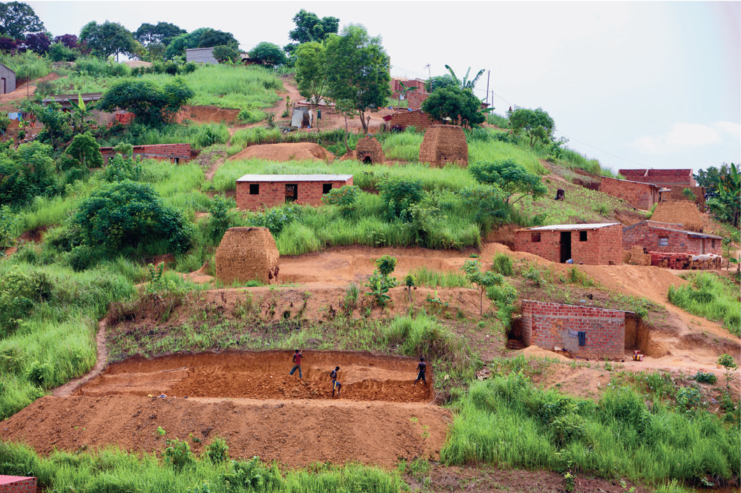 Imagem: Fotografia. Campo de terra com vegetação e árvores entre casas pequenas de tijolo e barrancos de terra.   Fim da imagem.