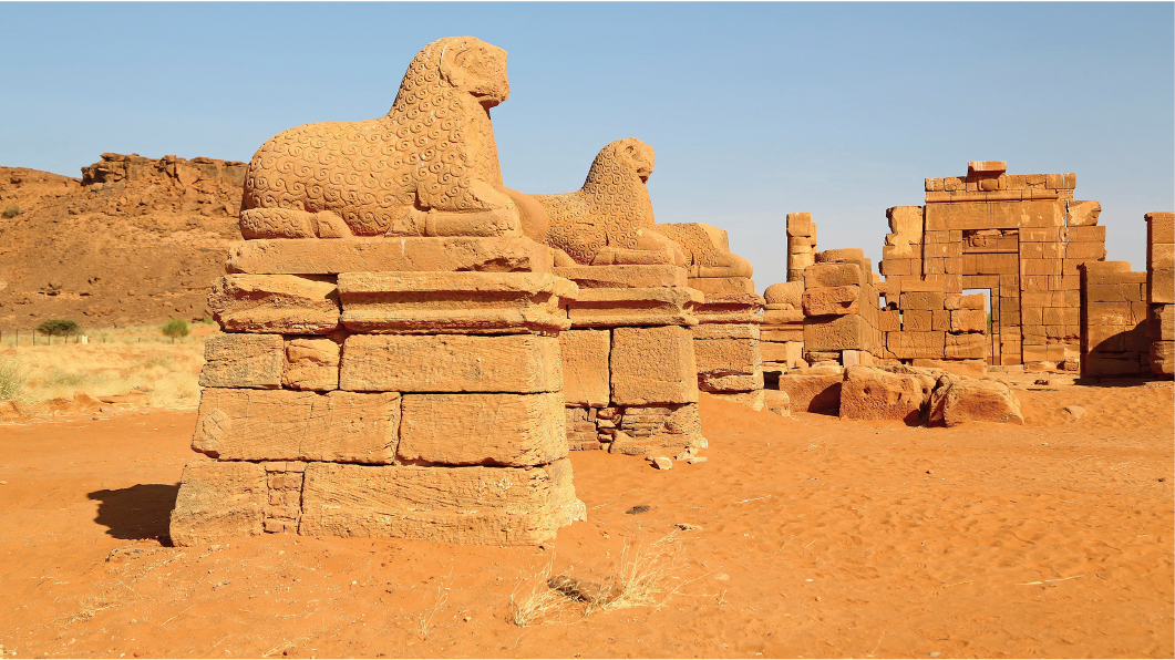 Imagem: Fotografia: Destaque de templo em ruínas com escultura de animais deitados em colunas baixas.  Fim da imagem.