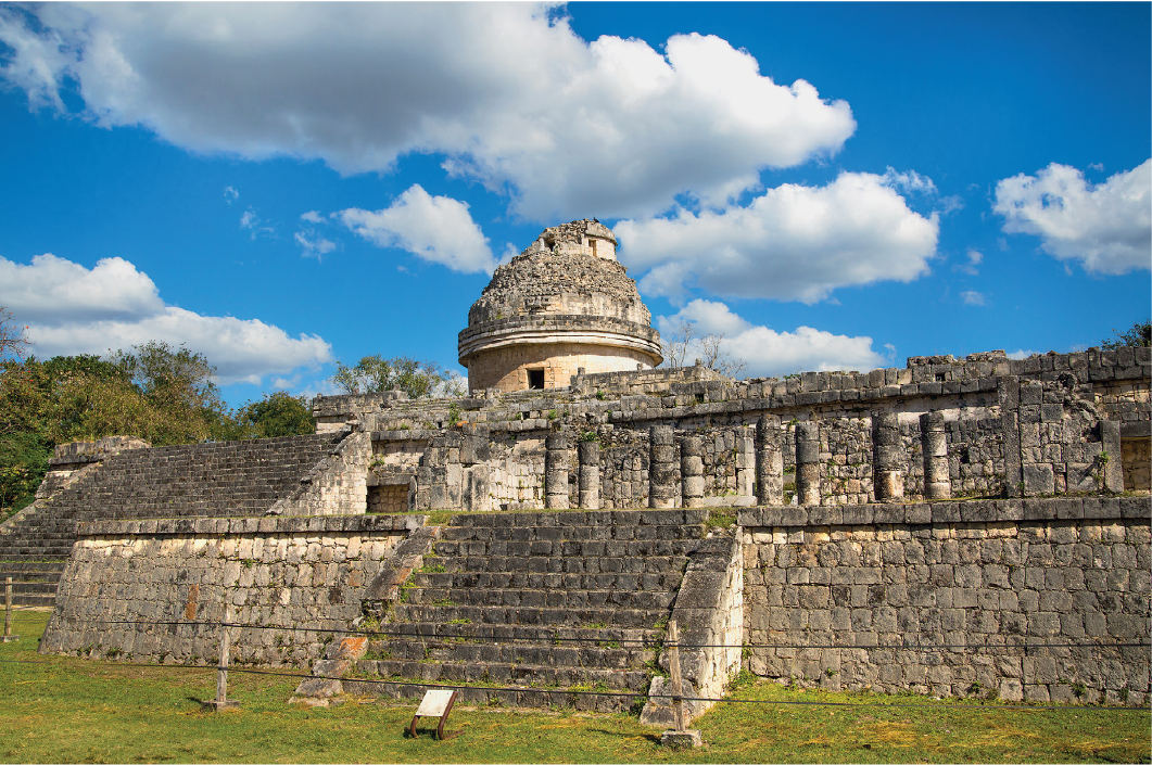 Imagem: Fotografia. Faixada de templo de pedras com escadas centrais e colunas de pedras arredondadas acopladas a um paredão de pedras. Ao fundo, uma estrutura cilíndrica com aberturas e telhado arredondado.   Fim da imagem.