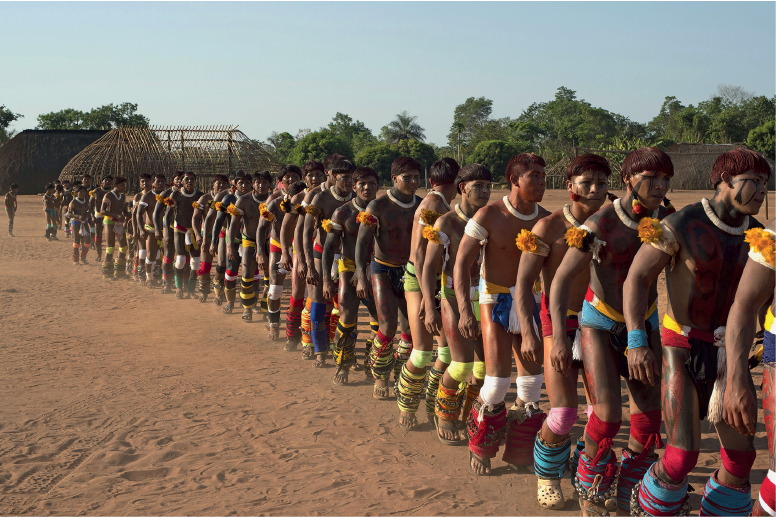 Imagem: Fotografia. Campo de areia com fila extensa de homens de rostos e corpo pintado com tanga colorida e adornos coloridos pelo corpo.   Fim da imagem.