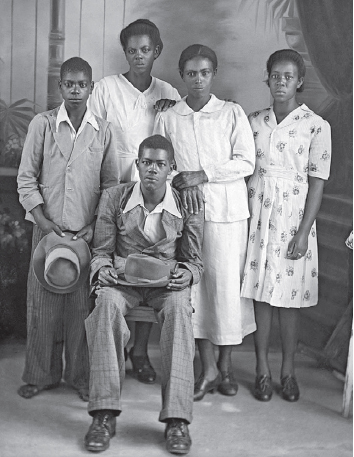 Imagem: Fotografia em preto e banco. Homem de cabelo curto, vestindo camisa, paletó e calça. Está sentado em uma cadeira, segurando um chapéu. Ao redor, menino de cabelo curto, vestindo paletó e calça, segurando um chapéu, três mulheres de cabelo preso, vestindo vestido até o joelho.  Fim da imagem.