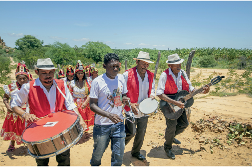 Imagem: Fotografia. Homens em campo aberto com vegetação baixa. Homens veste camiseta branca e colete vermelho, estão tocando instrumentos musicais. Atrás há mulheres de vestido longo branco e vermelho com chapéu vermelho na cabeça.  Fim da imagem.