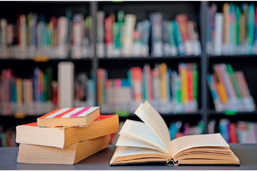 Imagem: Fotografia. Destaque de uma mesa com livros abertos. Ao fundo, há estantes com livros empilhados.  Fim da imagem.