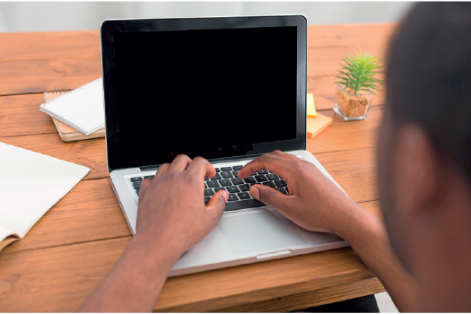 Imagem: Fotografia. Destaque de mãos de um homem digitando em um notebook apoiado sobre uma mesa de madeira. Ao redor há cadernos.  Fim da imagem.