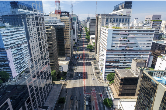 Imagem: Fotografia. Vista aérea de avenida dupla com três faixas cada lado. Ao redor da rua há prédios e árvores.   Fim da imagem.