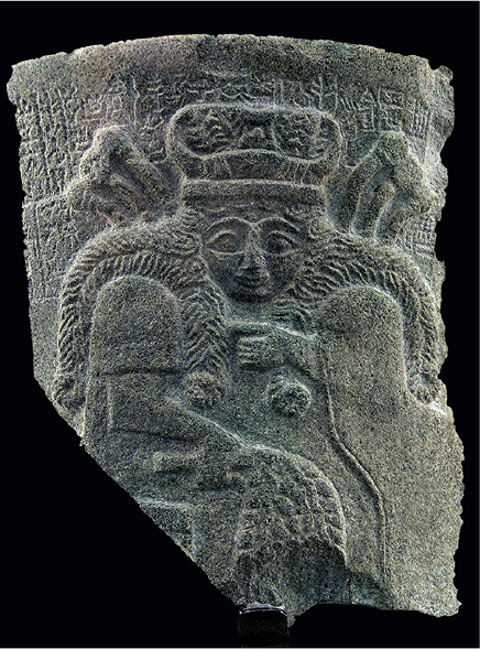 Imagem: Fotografia. Pedra retangular com pontas quebradas, no centro há uma mulher de coroa, cabelo longo cacheado, segurando um ramo.  Fim da imagem.