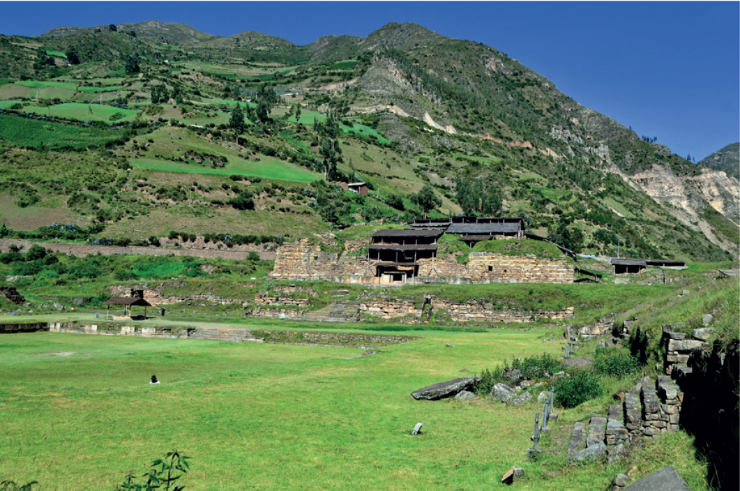 Imagem: Fotografia. Vista de campo aberto com morros e ruínas de sítio arqueológico com casas e construções. Ao fundo há montanhas com vegetação baixa e poucas árvores.   Fim da imagem.