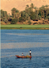 Imagem: Fotografia. Vista de canoa com duas pessoas sobre rio largo. Ao redor há vegetação pena margem e morros de areia ao fundo.   Fim da imagem.