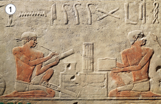 Imagem: Fotografia. Parede em alto-relevo com ilustração de homens ajoelhados trabalhando em papiros.  Fim da imagem.