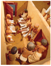 Imagem: Fotografia. Escultura de madeira com homens trabalhando em uma pequena sala com papéis e instrumentos de escrita.  Fim da imagem.
