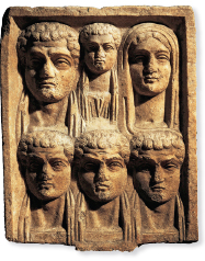 Imagem: Fotografia. Escultura retangular com o rosto de seis pessoas, duas mulheres e quatro homens.  Fim da imagem.