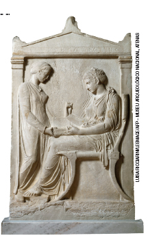Imagem: Fotografia. Escultura em alto-relevo de duas mulheres vestindo túnica. Uma mulher em pé segura uma caixa no colo de uma mulher sentada em um trono. Fim da imagem.