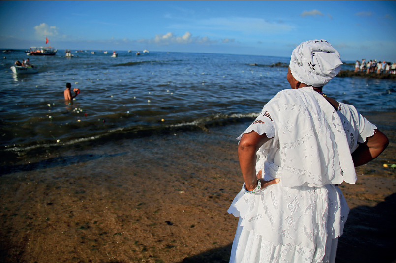 Imagem: Fotografia. Mulher de turbante e vestido longo branco, está na beira do mar.   Fim da imagem.
