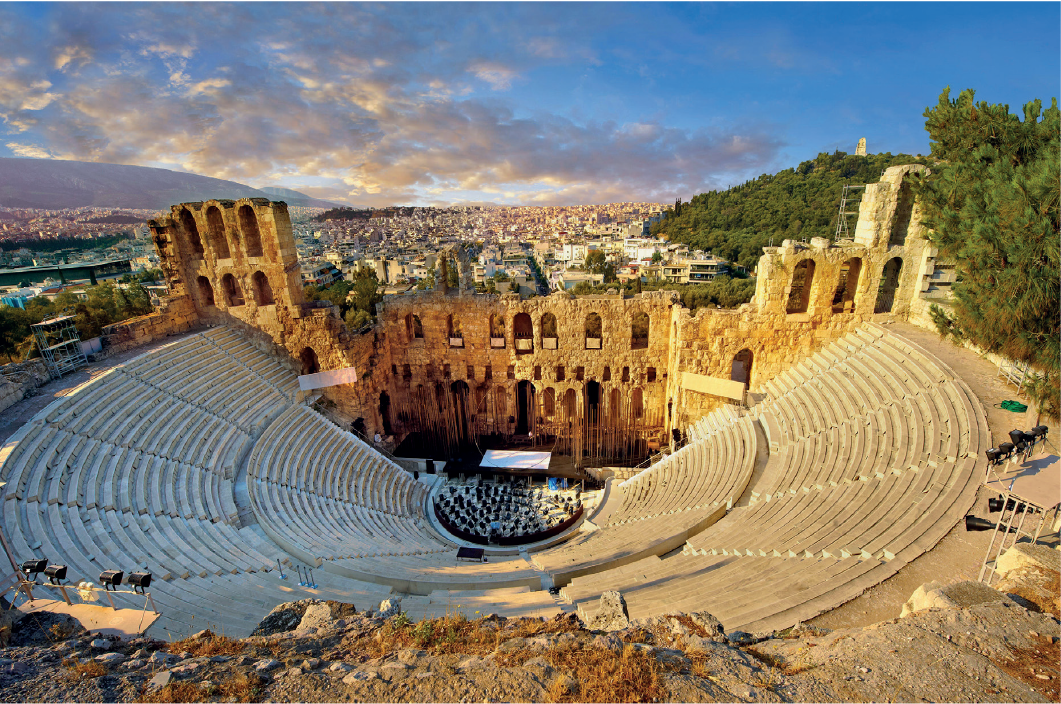 Imagem: Fotografia: Vista superior de arquibancada de pedra formando um semicírculo ao redor do palco, atrás do palco há ruinas de muros com janelas e arcos.   Fim da imagem.