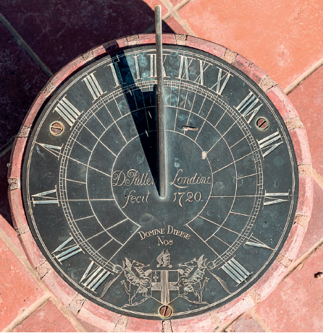 Imagem: Fotografia. Vista superior de relógio de sol preto com símbolos cinzas com números romanos ao redor e ponteiro retangular de pedra formando sombra pela estrutura.   Fim da imagem.