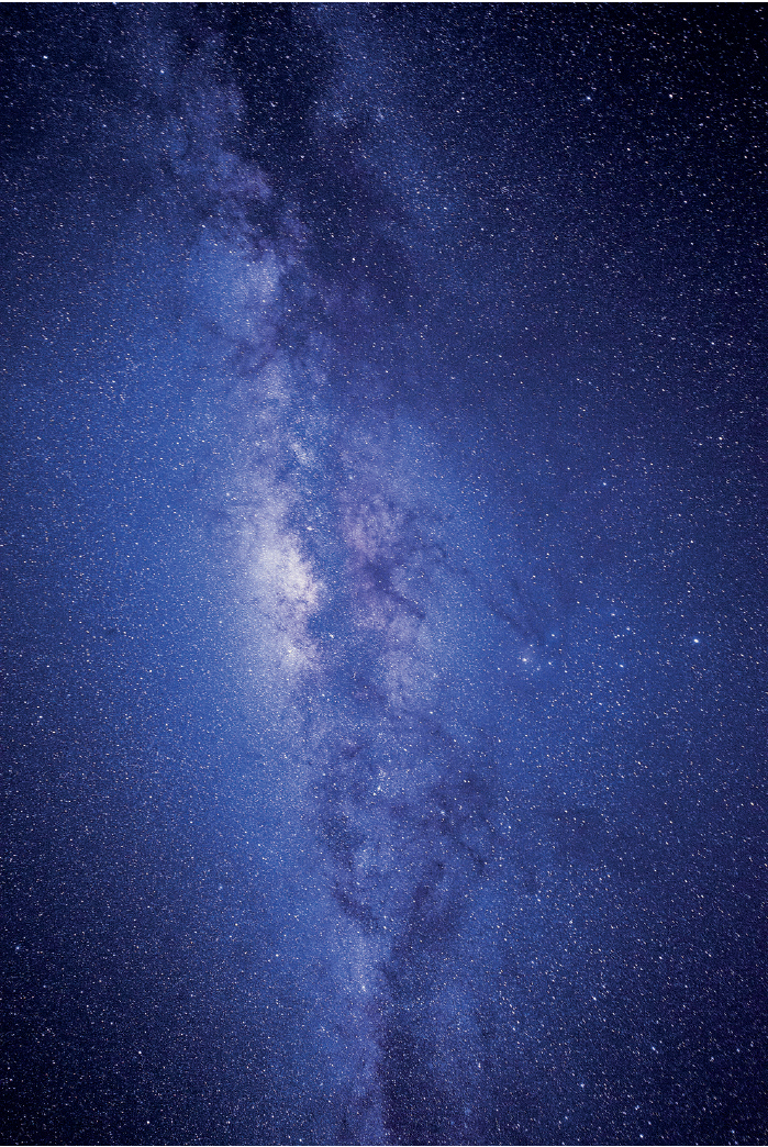 Imagem: Fotografia. Via láctea com estrelas em tons de azul e faixa clareada ao centro em tons de branco, azul e roxo.   Fim da imagem.
