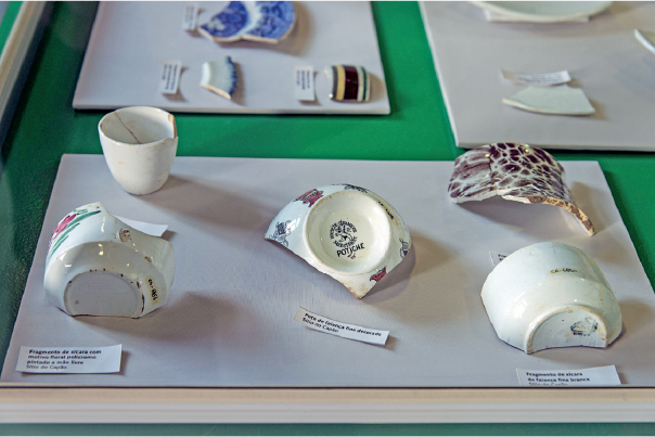 Imagem: Fotografia. Destaque de placas de informação com objetos quebrados e lascados de cerâmica pintada.   Fim da imagem.