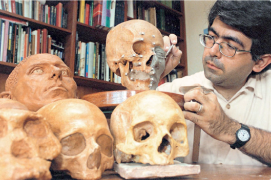 Imagem: Fotografia. Homem de cabelo curto preto e óculos de armação arredondada preta, vestindo camiseta bege. Ao lado há uma mesa com peças em forma de crânio. Ele está manuseando massa de reconstituição facial.   Fim da imagem.