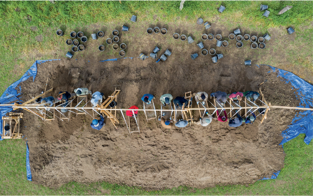 Imagem: Fotografia. Vista superior de homens trabalhando em escavação em campo aberto. Há sistema de madeira auxiliando áreas de estudo.   Fim da imagem.