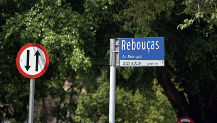 Imagem: Fotografia. Destaque de duas placas em rua. Uma placa circular com contorno vermelho e interior branco, com duas setas pretas verticais em direções opostas. Ao lado, placa retangular azul destacando nome da rua “Rebouças”.   Fim da imagem.