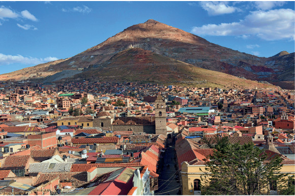 Imagem: Fotografia. Vista superior de casas e poucos prédios baixos. Em segundo plano há duas montanhas com vegetação baixa em tons de laranja e vermelho.  Fim da imagem.