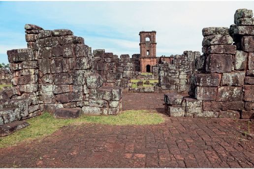 Imagem: Fotografia. Sítio arqueológico com ruinas de muros formados por pedras grande e largas.  Fim da imagem.