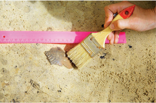 Imagem: Fotografia. Destaque de uma mão passando um pincel por peça de cerâmica coberta por areia. Ao lado, posicionado ao chão, há uma régua rosa.   Fim da imagem.