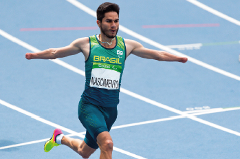 Imagem: Fotografia. Um homem, vestindo regata em tons de verde escrita BRASIL e uma placa de identificação escrito NASCIMENTO, corre com os braços abertos. No final de seus braços, mãos amputadas. Ao fundo, uma pista de corrida.  Fim da imagem.