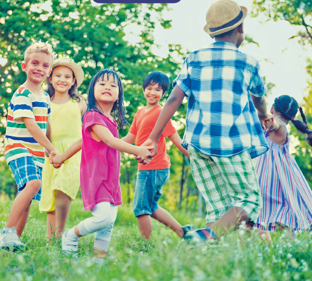 Imagem: Fotografia. Seis crianças sorrindo e de mãos dadas, formam um círculo. O ambiente é ensolarado e possui grama baixa. Ao fundo, árvores altas. Fim da imagem.