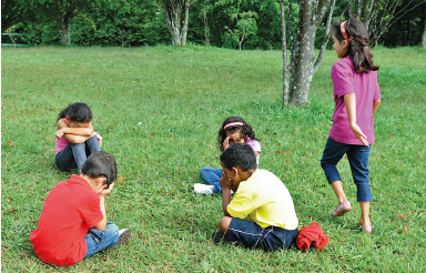 Imagem: Fotografia. Quatro crianças estão sentadas na grama, com os olhos tampados pelas mãos e as cabeças baixas. À direita das crianças, outra criança está de pé.  Fim da imagem.