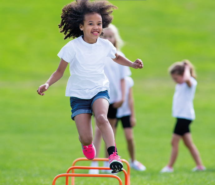 Imagem: Fotografia. Uma criança, com cabelo cacheado, camiseta branca, bermuda azul e tênis rosa, salta um obstáculo laranja fixado no chão. Ao fundo, crianças e grama.  Fim da imagem.