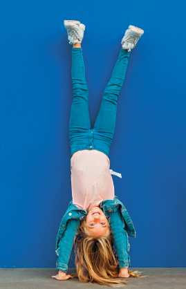 Imagem: Fotografia. Uma criança, de camisa branca e calça azul, está de cabeça para baixo. As mãos estão no chão e as pernas para cima. O fundo é azul.  Fim da imagem.