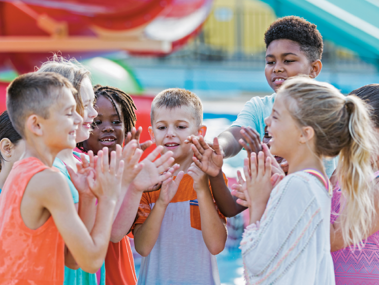 Imagem: Fotografia. Grupo de crianças com os braços dobrados e com as mãos na altura do queixo, batem palmas e sorriem.  Fim da imagem.