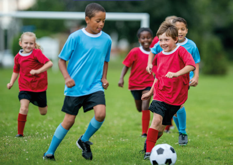 Imagem: Fotografia. Na frente, duas crianças vestindo camiseta, bermuda e meias altas, olham para uma bola de futebol no chão. Algumas estão de camisetas vermelhas e outras camisetas azuis. Ao fundo, outras crianças correm.  Fim da imagem.