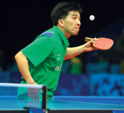 Imagem: Fotografia. Um homem vestindo camiseta verde, segura uma raquete pequena na mão esquerda e aponta na direção de uma bola branca. Na frente dele, há parte de uma mesa azul com uma rede no meio.  Fim da imagem.