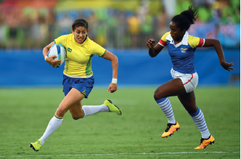 Imagem: Fotografia. À direita, há uma mulher, vestindo camiseta amarela e bermuda azul, está com os braços e pernas flexionados e segura uma bola oval branca no braço direito. À esquerda, há outra mulher, vestindo camiseta azul e bermuda branca, com os braços e pernas flexionados na direção da bola. O chão é de grama.  Fim da imagem.