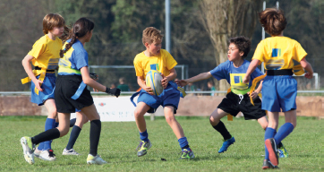 Imagem: Fotografia. No centro, uma criança com camisa amarela e bermuda azul, segura uma bola oval branca com as duas mãos. Ao redor dele, outras crianças estão olhando na direção da bola. Ao fundo, grama e árvores. Fim da imagem.