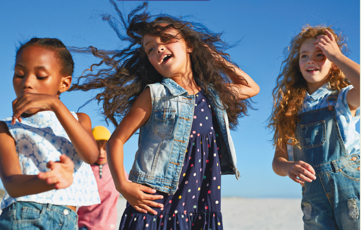 Imagem: Fotografia. À esquerda, uma menina com cabelo preso, vestindo regata branca, está com os braços inclinados. No centro, uma menina com cabelo longo e ondulado, usando vestido azul e jaqueta jeans, está com as mãos inclinadas na cintura e cabeça. À direita, uma menina com cabelo ruivo e longo, vestindo um macacão jeans e camiseta branca, está com a mão esquerda na cabeça. Fim da imagem.