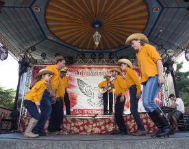 Imagem: Fotografia. No centro, duas fileiras de pessoas usando chapéu de palha, camiseta amarela, calça jeans e bota, estão com os joelhos dobrados. Ao fundo, em um palco, um homem está com chapéu de palha, camisa amarela, calça jeans e segura um violão.  Fim da imagem.