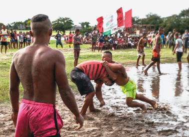 Imagem: Fotografia. À esquerda, um homem com tronco inclinado para frente e cabeça baixa. À direita, um homem inclinado para frente segura as costas do outro. O chão é de barro e, ao redor, há pessoas olhando.  Fim da imagem.