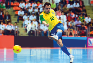 Imagem: Fotografia. No centro, um homem, vestindo camiseta amarela com o número 12 e bermuda azul, está com a perna esquerda levantada e olha para uma bola amarela de futsal. Ao fundo, pessoas sentadas na arquibancada. Fim da imagem.