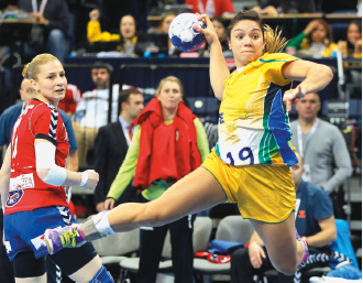 Imagem: Fotografia. No centro, uma mulher, vestindo camiseta e bermuda amarela, está com as sobrancelhas levantadas e a boca contraída. As pernas e braços estão flexionados e ela segura uma bola branca com a mão direita. Ao fundo, uma mulher, vestindo roupa vermelha, olha para a bola.  Fim da imagem.