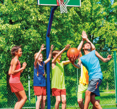 Imagem: Fotografia. No centro, um menino vestindo camiseta azul e bermuda cinza, está com os braços dobrados e segura uma bola de basquete com a mão direita. Atrás dele, quatro crianças estão com os braços levantados e olham para a bola. Ao fundo, uma cesta de basquete. Fim da imagem.