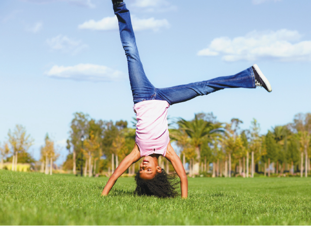 Imagem: Fotografia. Uma criança vestindo regata rosa e calça jeans está de ponta cabeça, com as mãos apoiadas no chão gramado e as pernas para cima.  Fim da imagem.