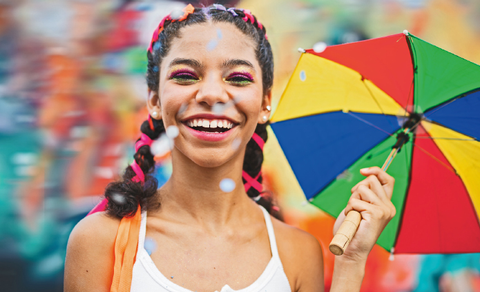 Imagem: Fotografia. Destaque para o rosto de uma menina com cabelo preso em maria-chiquinhas amarrado com fitas coloridas e sorrindo. Com a mão esquerda, ela segura um guarda-chuva pequeno e colorido.  Fim da imagem.